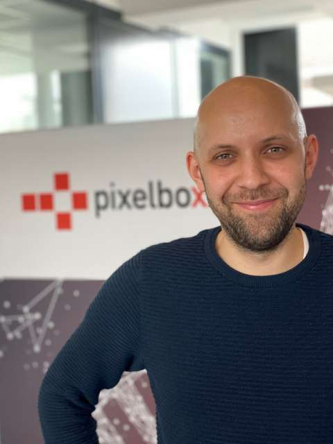 Ein Portraitbild eines lächelnden jungen Mannes mit Dreitagebart und Glatze vor einem Pixelboxx Aufsteller.