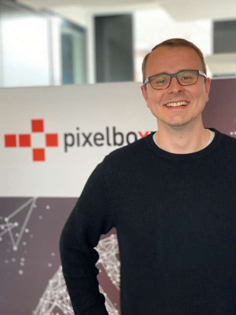 Ein junger Mann mit dunklem Pullover und Brille steht lächelnd vor einem Pixelboxx Aufsteller